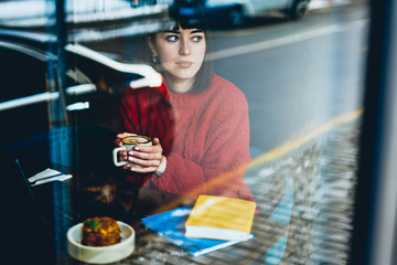 Caucasian brunette hipster girl thinking pondering about learning spending time for sweet lunch with caffeine beverage, thoughtful female student holding cup of cappuccino and looking at window