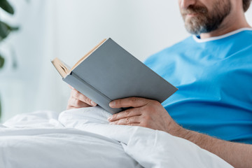 cropped view of patient in medical gown reading book in hospital