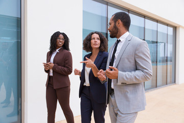 Fototapeta premium Positive multiethnic business colleagues walking near office building. Man and women wearing formal suits, going past outdoor wall, talking to each other. Corporate friends concept