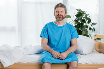 smiling patient in medical gown sitting on bed and looking at camera in hospital
