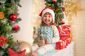little girl in pajamas and Santa hat drinking cocoa milk! Christmas atmosphere surrounds. boxes of gifts tied with satin ribbons. the Christmas tree garland is decorated with dried oranges and balls