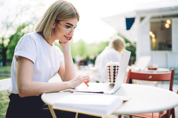 Glad female student in glasses using laptop in cafe