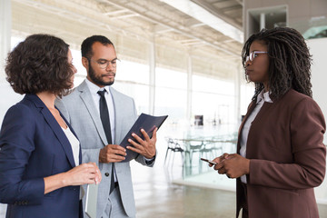 Business partners discussing contract terms. Business man and women standing in modern office hallway, holding documents and digital devices, discussing project. Business meeting concept