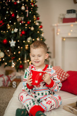 A little boy in Christmas pajamas sits near a Christmas tree and holds a mug of cocoa with marshmallows. Christmas. New Year.