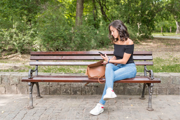 One lonely beautiful girl sitting alone and depressed on the bench in the park feeling abandoned and betrayed from her boyfriend when he suddenly break up with her by text message on the phone