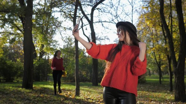 femininity, a young attractive girl cheerfully runs to meet her pretty girlfriend in a hat and take a selfie on the phone with fallen leaves toss him in a beautiful park