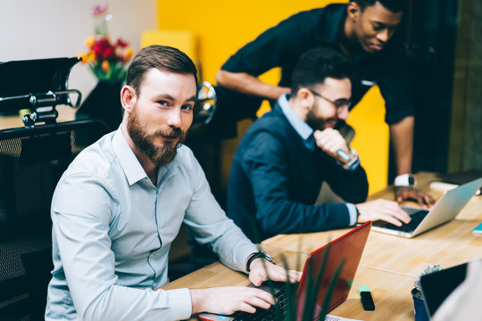 Group of young it professionals developing program code for antivirus on laptop computer enjoying collaboration indoors, portrait of successful man looking at camera while colleagues talking near