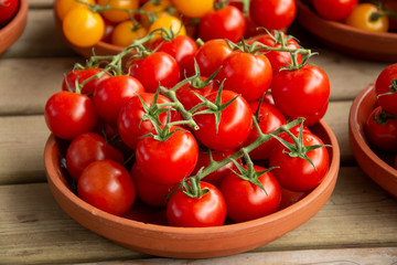 A bunch of red tomatoes in a clay plate surrounded by other tomatoes plate over a wooden table