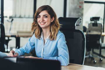 Portrait of prosperous beautiful young woman dressed in formal wear smiling at camera while writing down plan organization of working process.Cheerful businesswoman making notes sitting at table