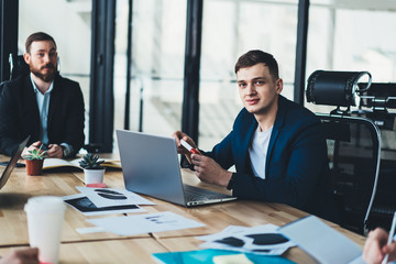 Portrait of successful young male entrepreneur in formal wear sitting at table with laptop computer during business meeting with colleagues in modern office.Traders looking at camera during briefing