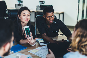 Young woman holding smartphone and showing app to colleague sitting at meeting table during briefing in office.Multicultural students discussing blog using mobile phone and wireless 4G internet