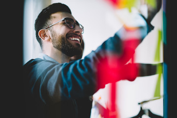 Cheerful young man in optical eyeglasses laughing while looking at stickers with notes and learning new words during lesson.Positive hipster guy smiling while reading information on colorful papers - obrazy, fototapety, plakaty