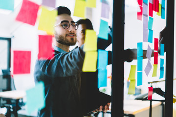 Diverse team of skilled young people learning foreign words from colorful stickers glued on glass wall during collaborative process in office interior.Multicultural students using paper sticky notes