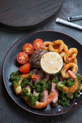 Surf and turf with marbled beef steaks, shrimps, calamari rings, cherry tomatoes and chimichurri served on a black plate, close-up