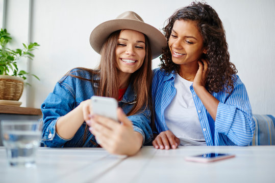 Smiling multiracial female friends viewing  photos on smartphone and share in social networks resting in cafe,cheerful hipster girl watching funny video on cellular enjoying free time together
