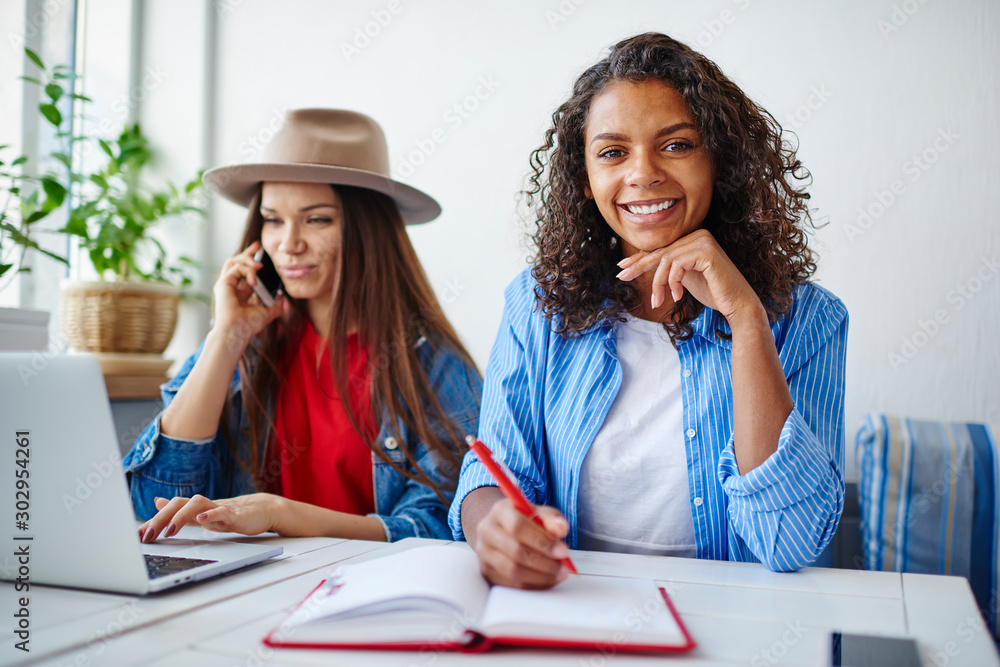 Poster portrait of cheerful dark skinned girl doing homework with her best friend talking on smartphone on 
