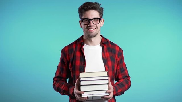 European student in red plaid shirt on blue background in studio holds stack of university books from library. Guy smiles, he is happy to graduate.