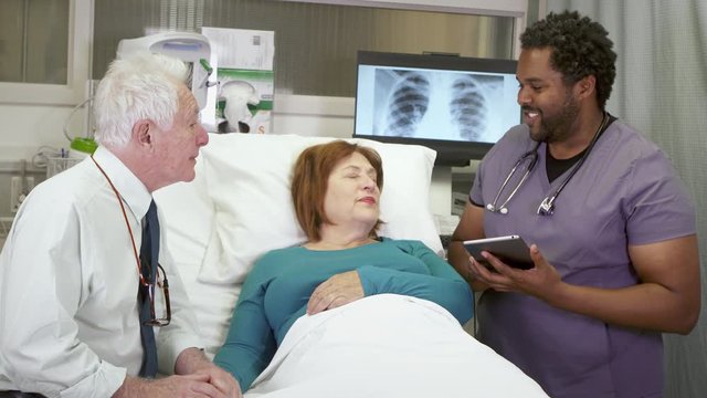 Modern Technology In A Hospital. A Black Male Nurse Uses A Mobile Tablet Computer To Deliver Good News To An Elderly Caucasian Couple Bedside At The Hospital.