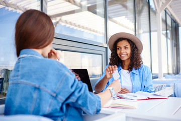 Cheerful african american girl happy about taking visit card from her best friend during meeting in cafe,female hipster advising best service to her dark skinned colleague giving visit card .