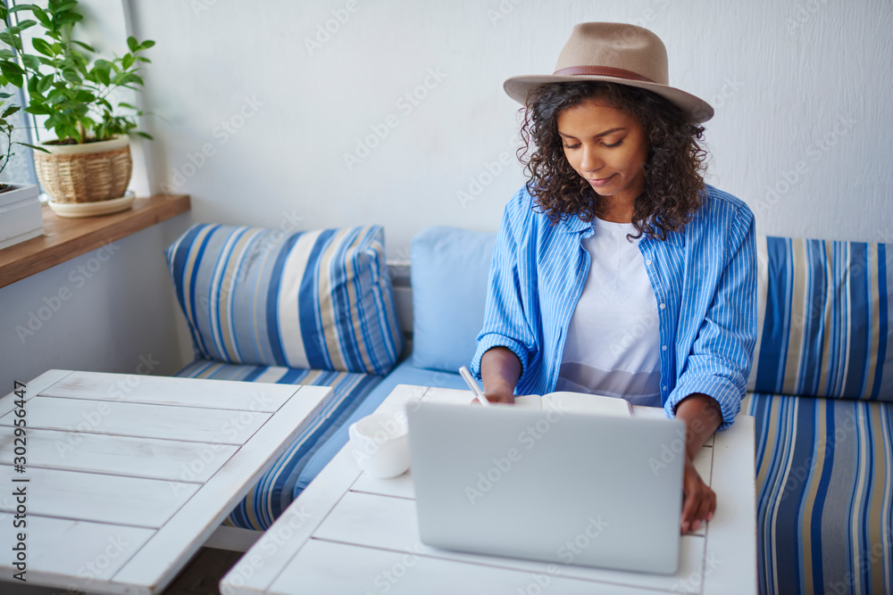 Poster dark skinned female student writing report for homework using laptop computer for browsing informati