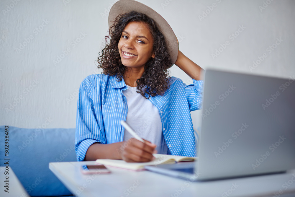 Poster clever african american hipster girl looking aat camera while doing homework task using laptop compu
