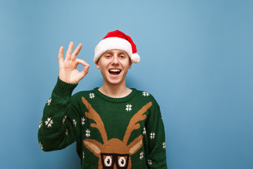 Cheerful guy in green christmas sweater and christmas hat stands on blue background, shows OK gesture, looks into camera and smiles. Joyful young man showing thumbs up gesture OK