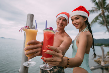 Young couple with cocktails in Christmas caps on the ocean on a tropical island.