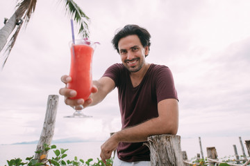 A young man stands by the sea on a wooden bridge with a cocktail.