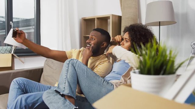 moving, repair and real estate concept - happy african american couple with cardboard boxes sitting on sofa and taking selfie by smartphone at new home
