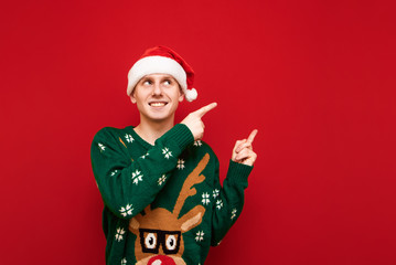 Smiling young christmas man in santa hat and warm slit sweater looking and pointing to the copy...