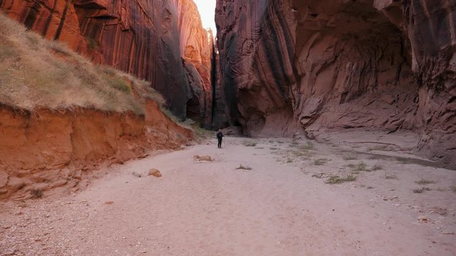 Hiker Stands At The Bottom Of A Deep Slot Canyon With High Red Stone Walls
