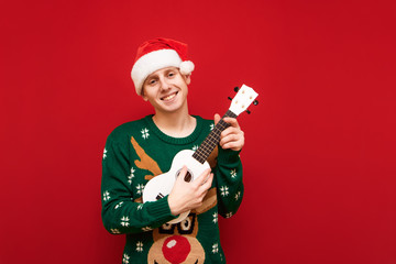 Happy young man in christmas clothes stands on red background with ukulele in hands, looks into camera and smiles, wears santa hat and green sweater. Isolated. Copy space - Powered by Adobe