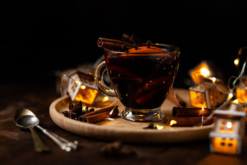 Christmas delicious hot mulled wine with orange, apple, cinnamon, clove, cardamom and anise on wooden plate. Warm beverage for cold winter days. Brights lights create festive mood. Dark background