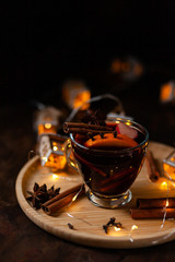 Christmas delicious hot mulled wine with orange, apple, cinnamon, clove, cardamom and anise on wooden plate. Warm beverage for cold winter days. Brights lights create festive mood. Dark background