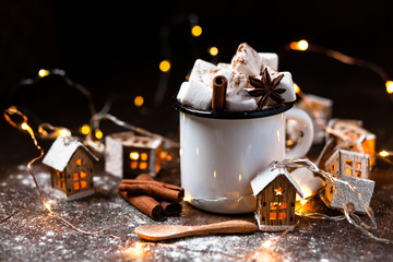 Winter hot drink: hot chocolate with marshmallows, cinnamon and anise. Christmas lights in the shape of houses as decor. Decorative snow, dark wooden background. Festive mood, cozy home atmosphere