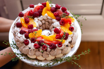 Young girl is holding delicious homemade Pavlova cake decorated with fresh oranges, raspberries and thyme. Traditional french dessert, sweet but light