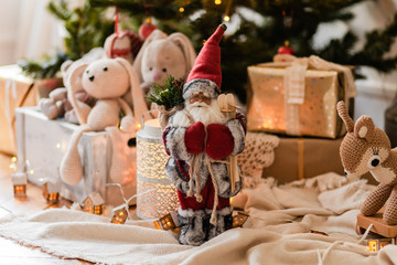 Christmas decor, rustic style. Santa Claus toy in red clothes, lamp and plush rabbits on background. Festive mood, cozy home atmosphere, family holiday  time. Green fir-tree with lamps on background