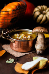Delicious hot porcini soup in copper pan. Tasty seasonal autumn meal. Fresh mashrooms illustrate ingredients. Concept of homemade food. Closeup, dark wooden background. Pumpkin on the back