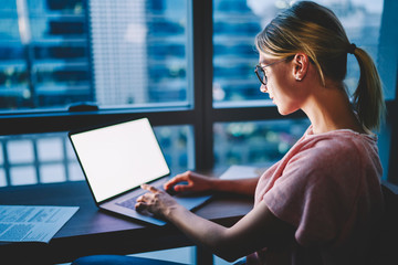 Young woman in eyewear typing text post for uploading on internet website on netbook with blank...