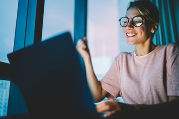 Accomplished student in eyewear feeling happy to compete course work late at night on laptop...
