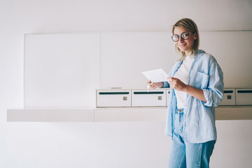 Cheerful caucasian blonde woman looking at camera holding white envelope from postal delivery, portrait of smiling hipster girl in eyewear satisfied with getting mail from friend in metal letterboxes