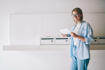 Cheerful caucasian blonde woman looking at camera holding white envelope from postal delivery, portrait of smiling hipster girl in eyewear satisfied with getting mail from friend in metal letterboxes