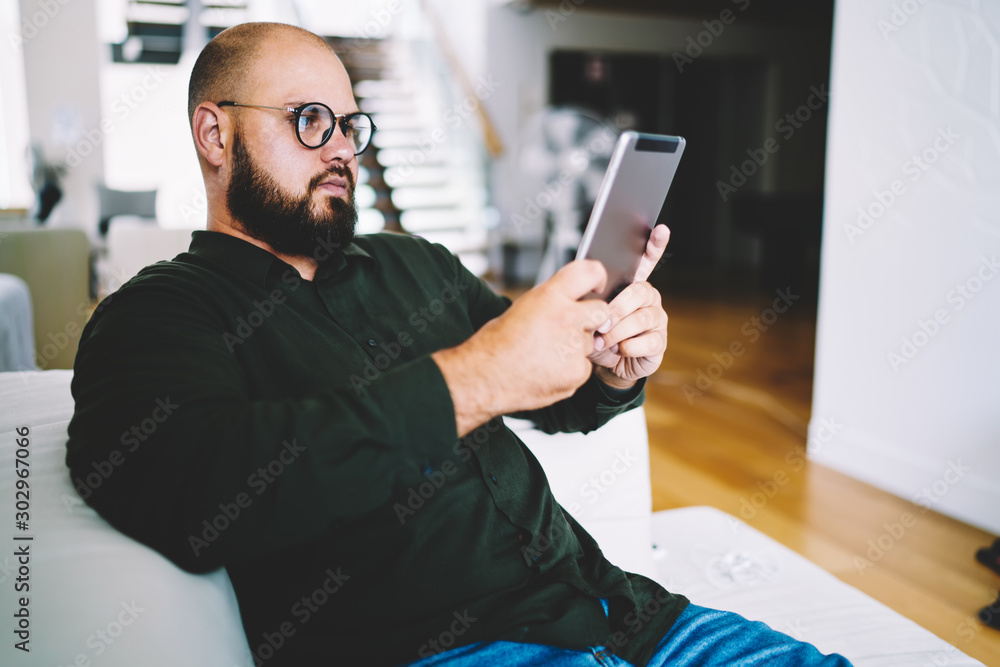 Poster concentrated casual dressed bearded man in eyewear reading e-book resting at home interior.male blog