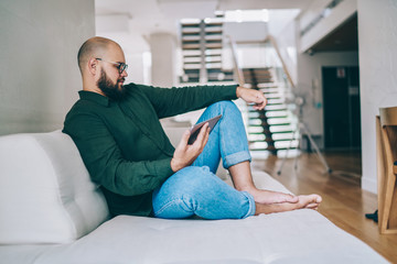 Fototapeta premium Positive hipster guy watching video satisfied with internet connection at home interior, caucasian male reading income message via digital tablet spending free time at comfortable couch in apartment
