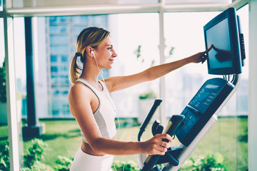 Young beautiful happy female person enjoying making exercises on bike for supporting healthy lifestyle and fit body. Pretty positive woman listening to music while training on cross trainer in gym