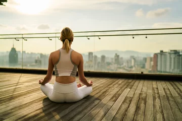 Fotobehang Back view of slim sports women care about body and health in megalopolis training yoga on rooftop of skyscraper, girl in track suit sitting in lotus pose feeling zen in city relaxation concept. © BullRun