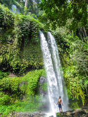 A man in swimsuit standing under two levelled waterfall in Lombok, Indonesia. Tiu Kelep Waterfall is surrounded by lush green plants from each side. Long and powerful waterfall. Beauty of the nature.