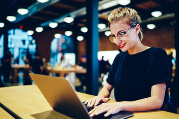 Young gorgeous smiling skilled designer in cool spectacles keyboarding email message for customer via laptop connected to 4G.Charming cheerful hipster girl sending funny messages to friends online
