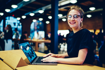 Half length portrait of charming emotional female blogger in trendy eyewear spending time sitting in coffee shop.Cute hipster girl sincerely laughing while looking at camera and installing application