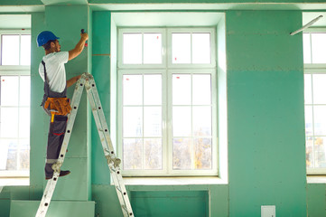 A builder standing on a ladder installs drywall at a construction site